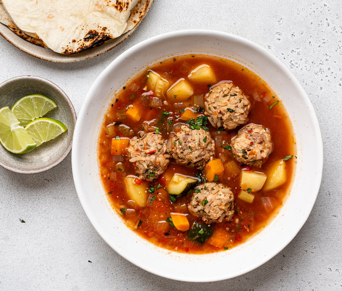 Albondigas Soup served in white bowl with a spoon and tortillas and lime wedges on the side.