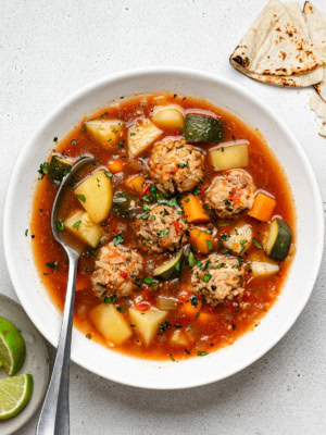 Albondigas Soup served in white bowl with a spoon and tortillas and lime wedges on the side.