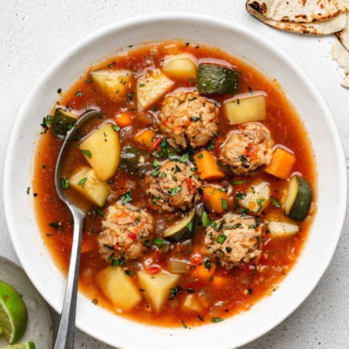 Albondigas Soup served in white bowl with a spoon and tortillas and lime wedges on the side.