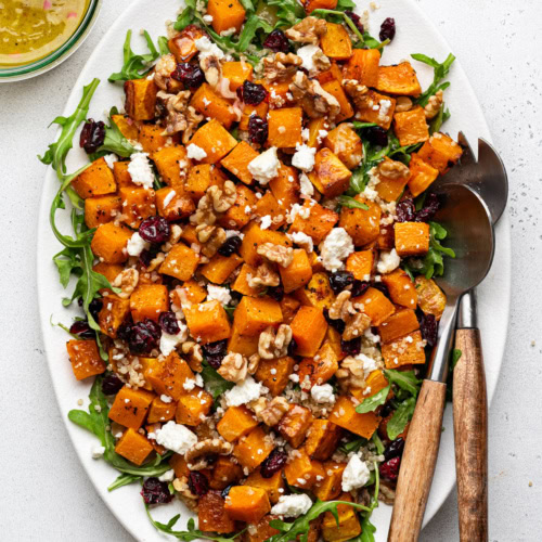 Butternut squash quinoa salad served on white platter with salad tongs and dressing in a jar on the side