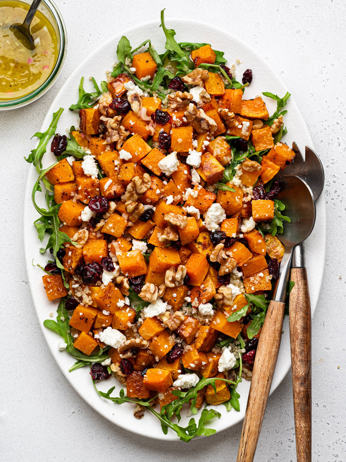 Butternut squash quinoa salad served on white platter with salad tongs and dressing in a jar on the side