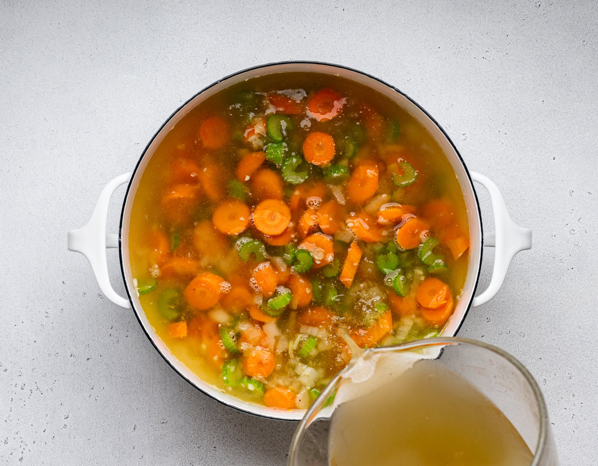 pouring broth over sautรฉed veggies in white soup pot