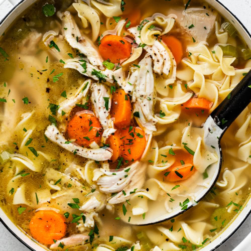 close-up of cooked turkey noodle soup in large white soup pot with a large spoon scooping out soup