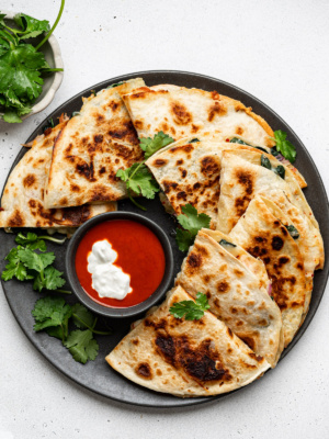 Quesadilla triangles served on a black plate, garnished with cilantro and a bowl of hot sauce and sour cream