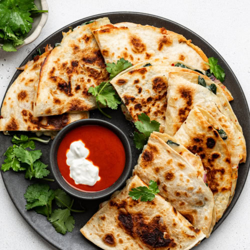 Quesadilla triangles served on a black plate, garnished with cilantro and a bowl of hot sauce and sour cream