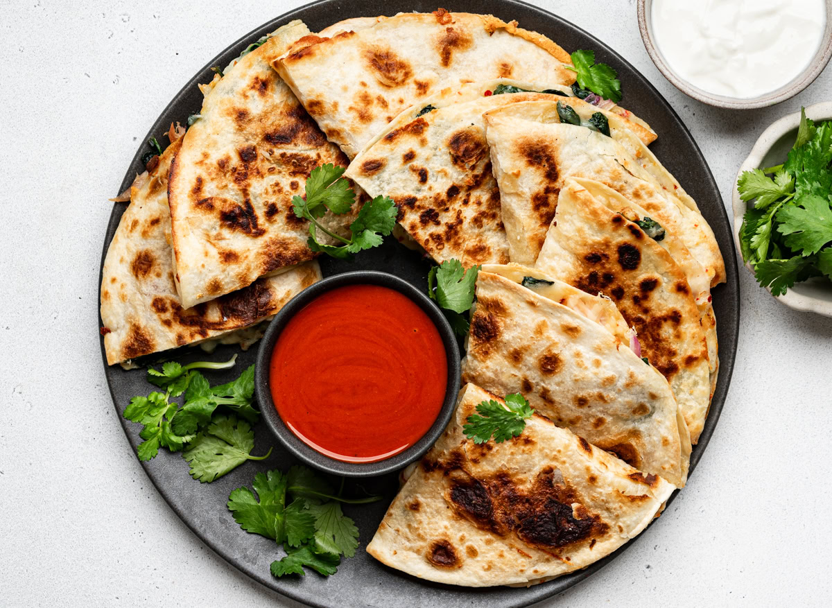 Quesadilla triangles served on a black plate, garnished with cilantro with bowls of hot sauce and sour cream on the side