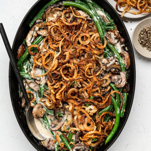 baked green bean casserole in oval black baking dish with crispy onions and black pepper in bowls on the side