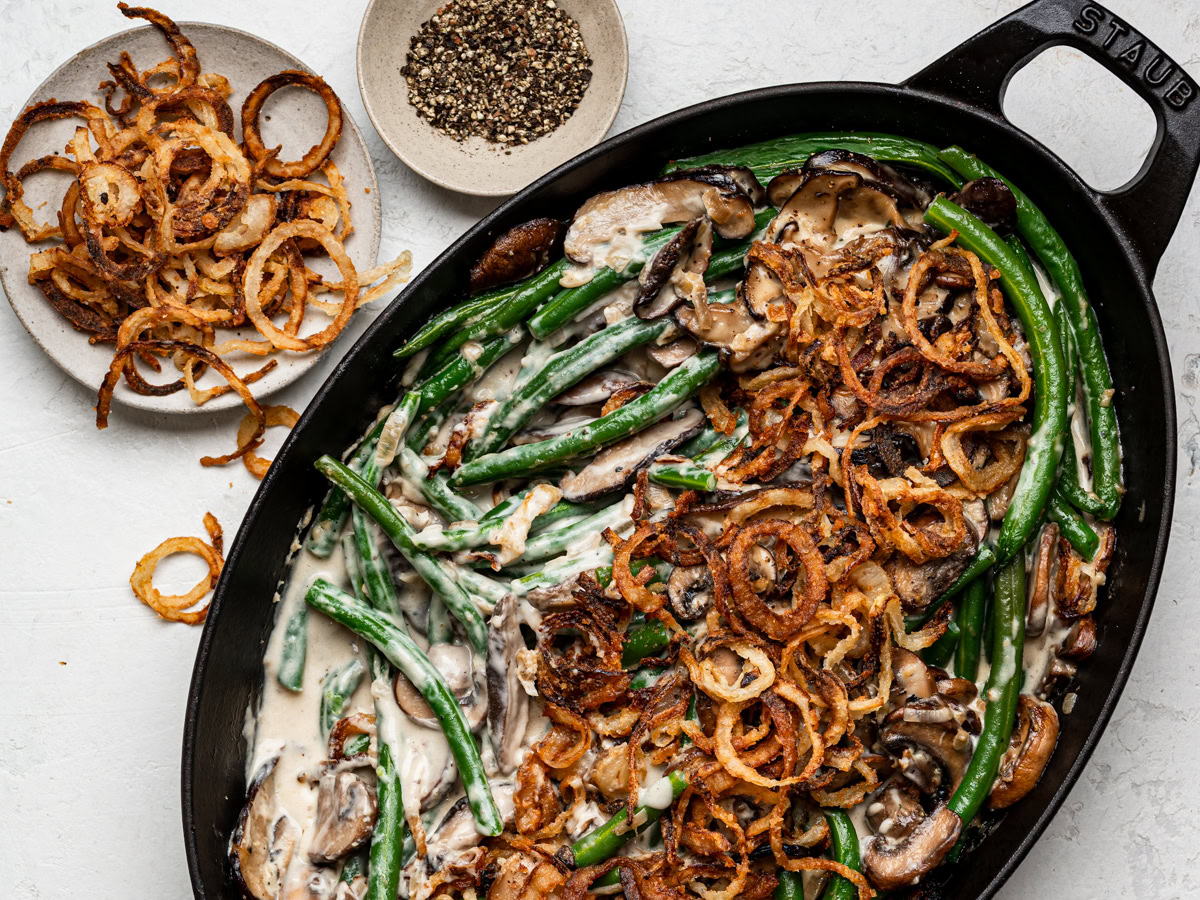 saucy green bean casserole from scratch in a black, oval baking dish with crispy onions and black pepper in bowls on the side