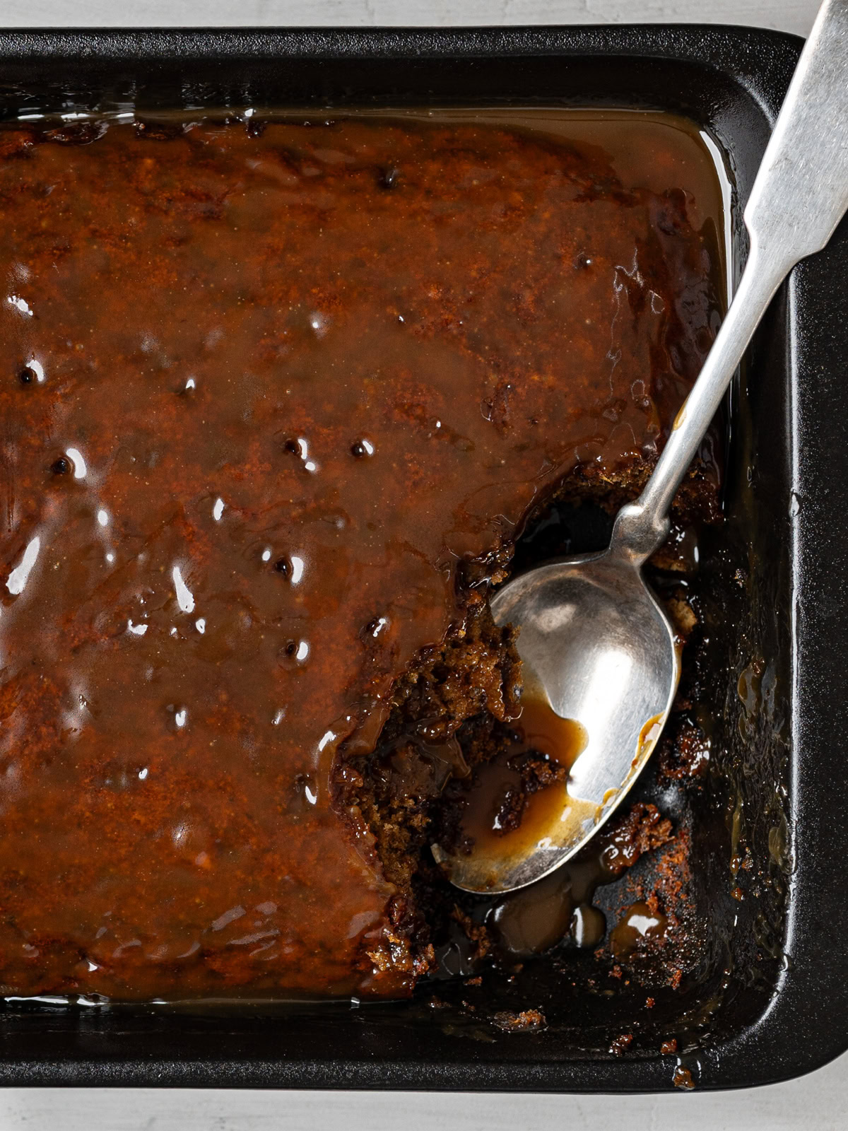 close up of baked sticky toffee cake in baking dish with some pieces scooped out and a silver spoon