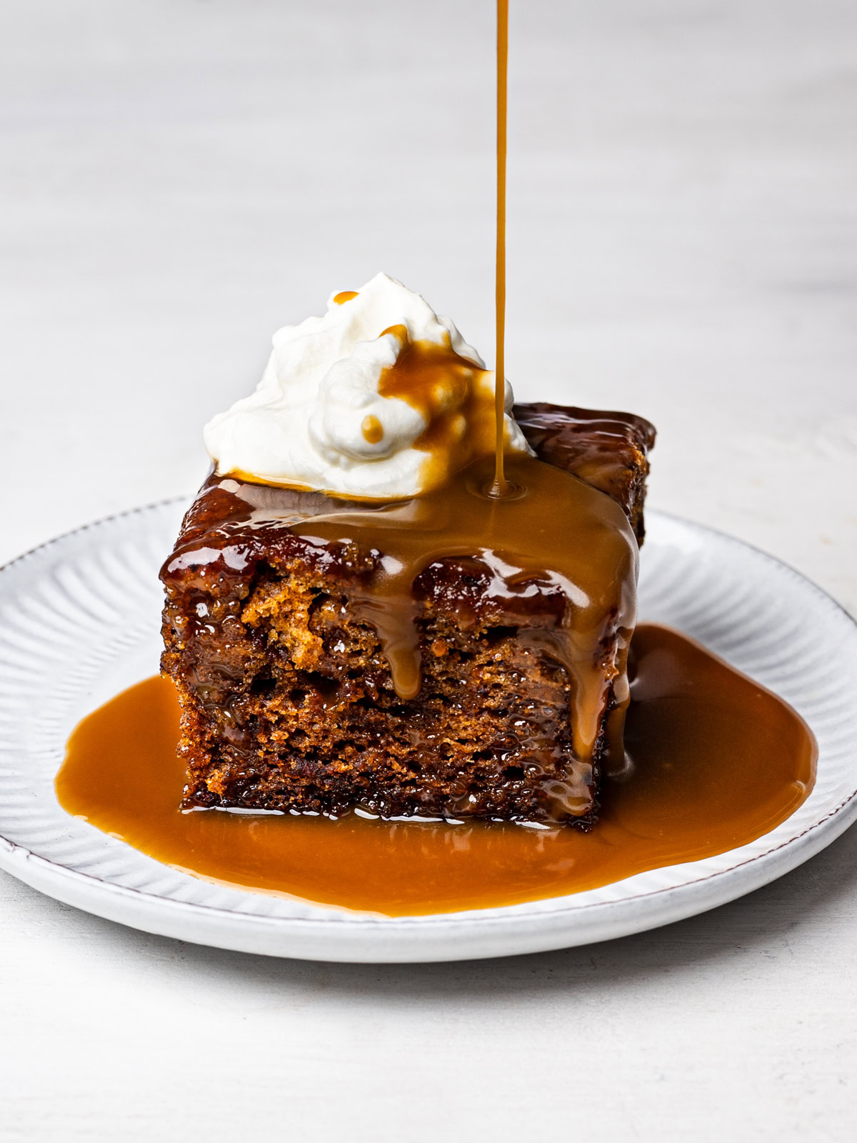 a square piece of sticky toffee date pudding served on a white small plate, topped with a dollop of whipped cream and drizzle of toffee sauce being poured over top