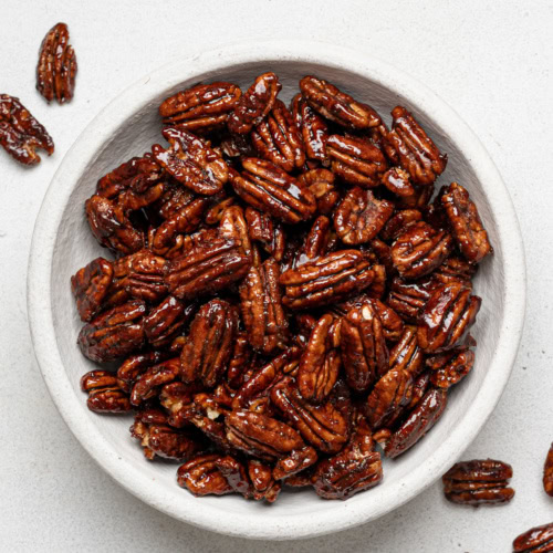 candied pecans served in white bowl with a few pecans fallen on the background