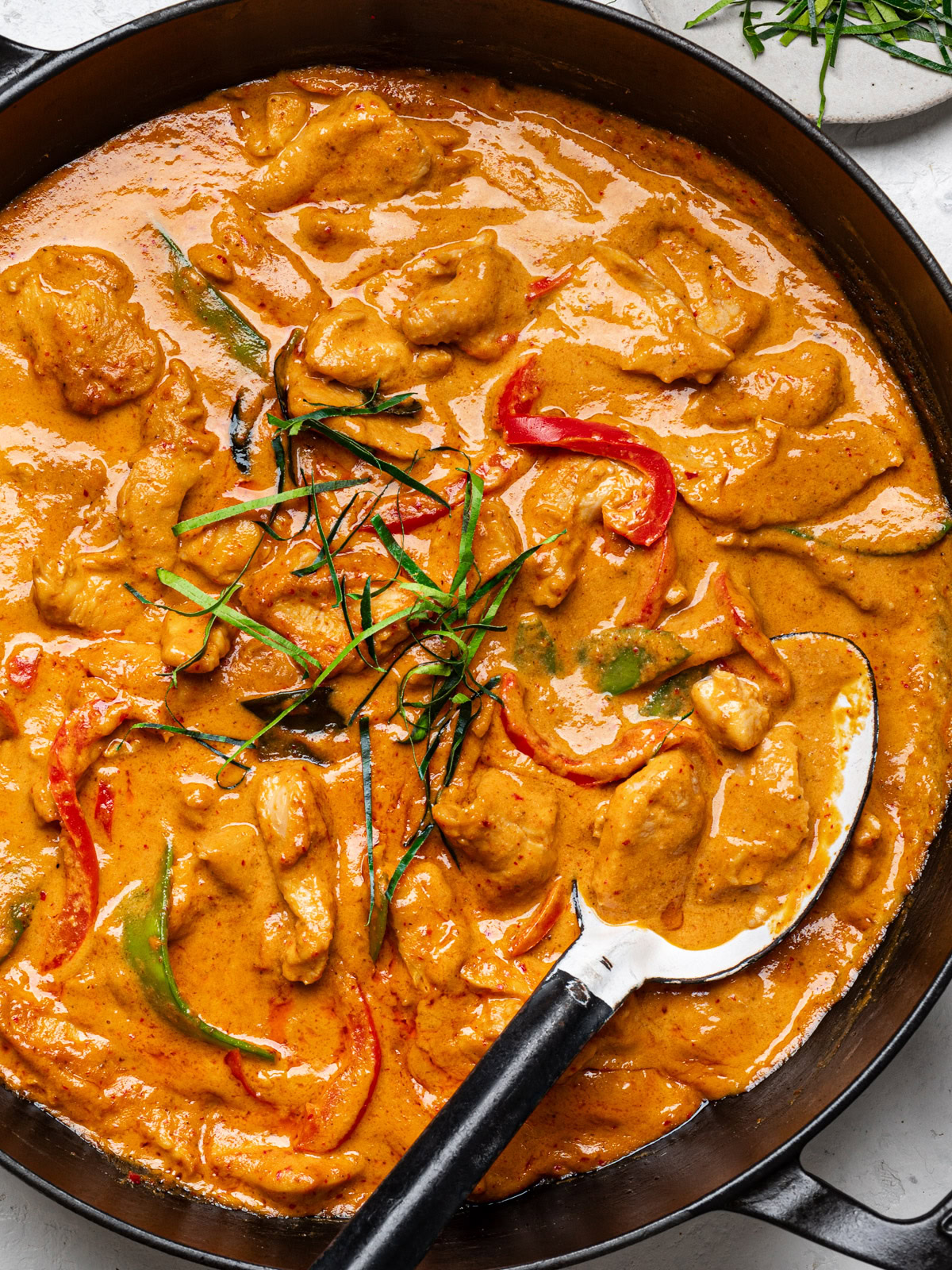 close up of finished panang chicken curry in black skillet garnished with sliced kaffir lime leaves and a large spoon scooping some of the curry