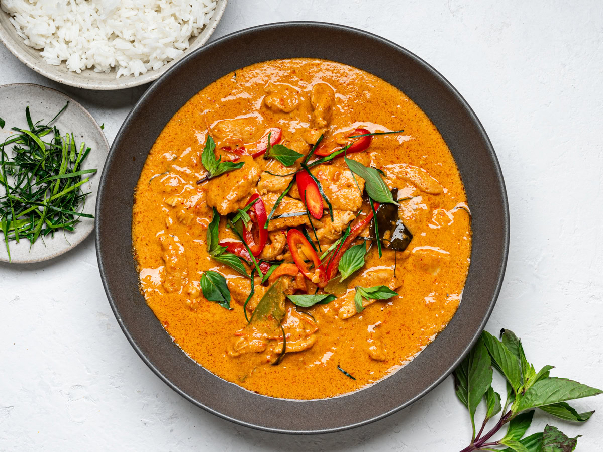 Panang chicken curry in a black bowl with white rice in a bowl on the side and another small bowl with sliced kaffir lime leaves