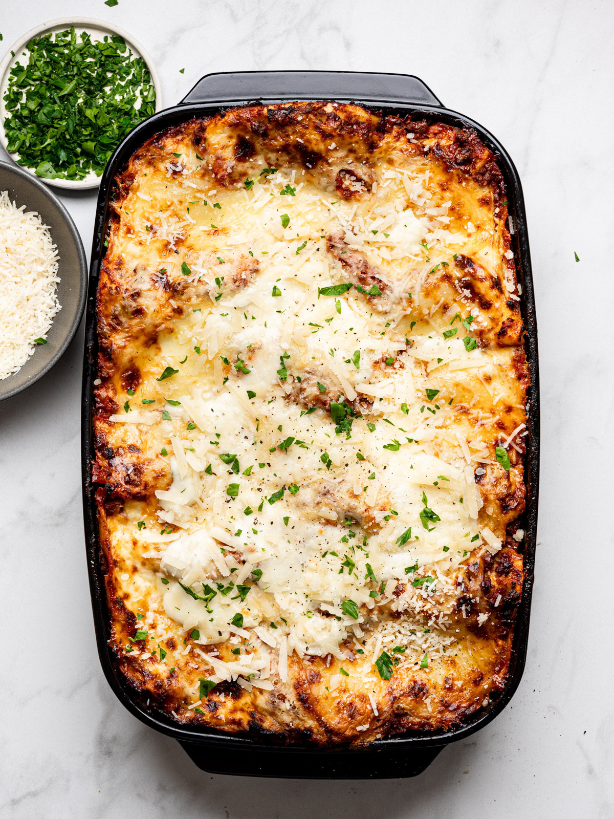 baked lasagna al forno in a black baking dish with chopped parsley in a small bowl on the side and bowl with parmesan cheese