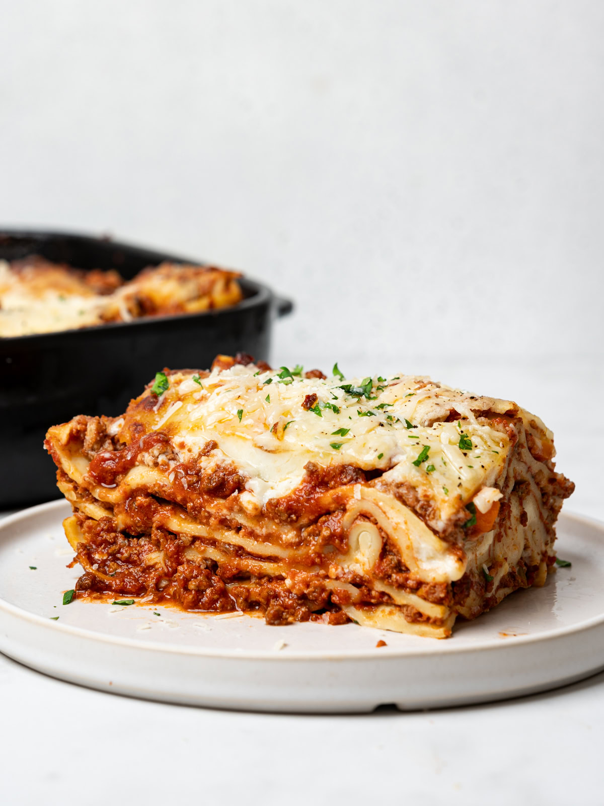 side view of a sliced serving of lasagna al forno on small white plate and black casserole dish in the background