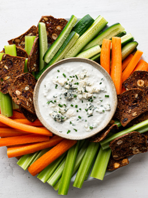 Blue cheese dip in bowl garnished with chopped chives and served with chopped vegetables and crackers on a round plate