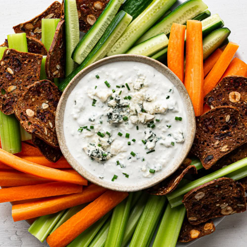 Blue cheese dip in bowl garnished with chopped chives and served with chopped vegetables and crackers on a round plate