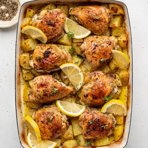 baked Greek chicken and potatoes in a white casserole dish garnished with lemon slices and fresh oregano.