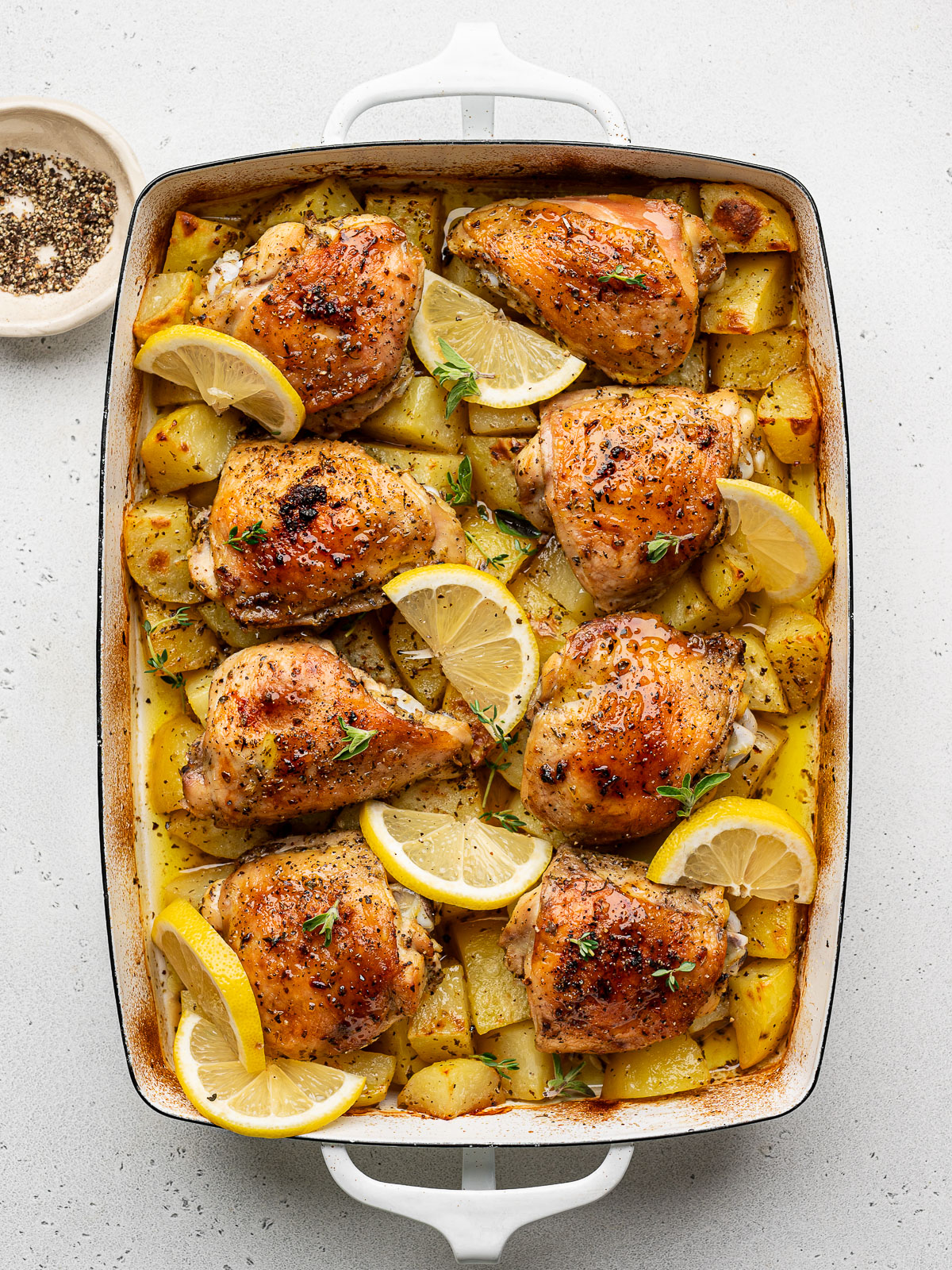 baked Greek chicken and potatoes in a white casserole dish garnished with lemon slices and fresh oregano.