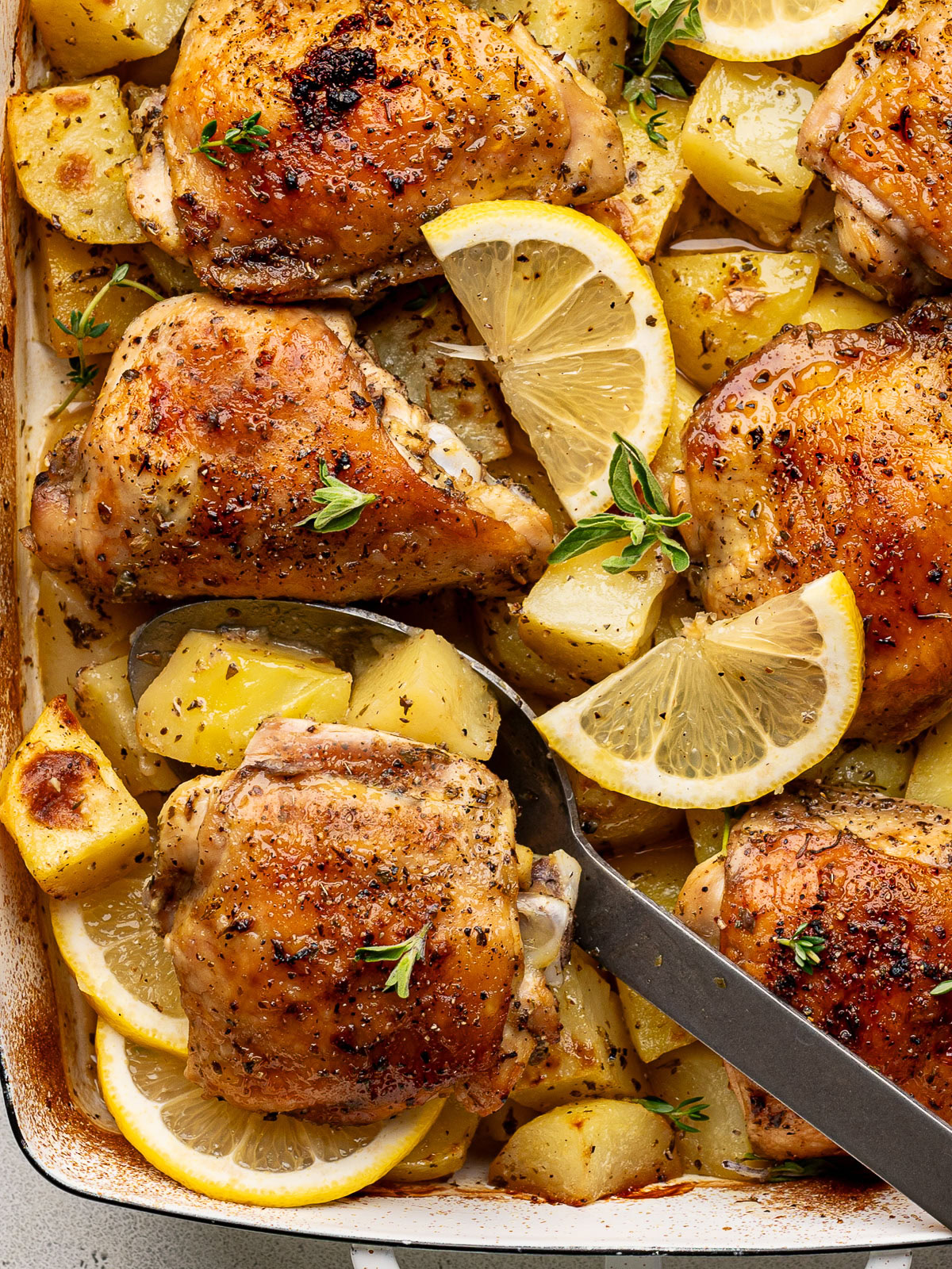 close up of baked Greek chicken and potatoes in a white casserole dish garnished with lemon slices and fresh oregano and a large serving spoon scooping out a piece of chicken.