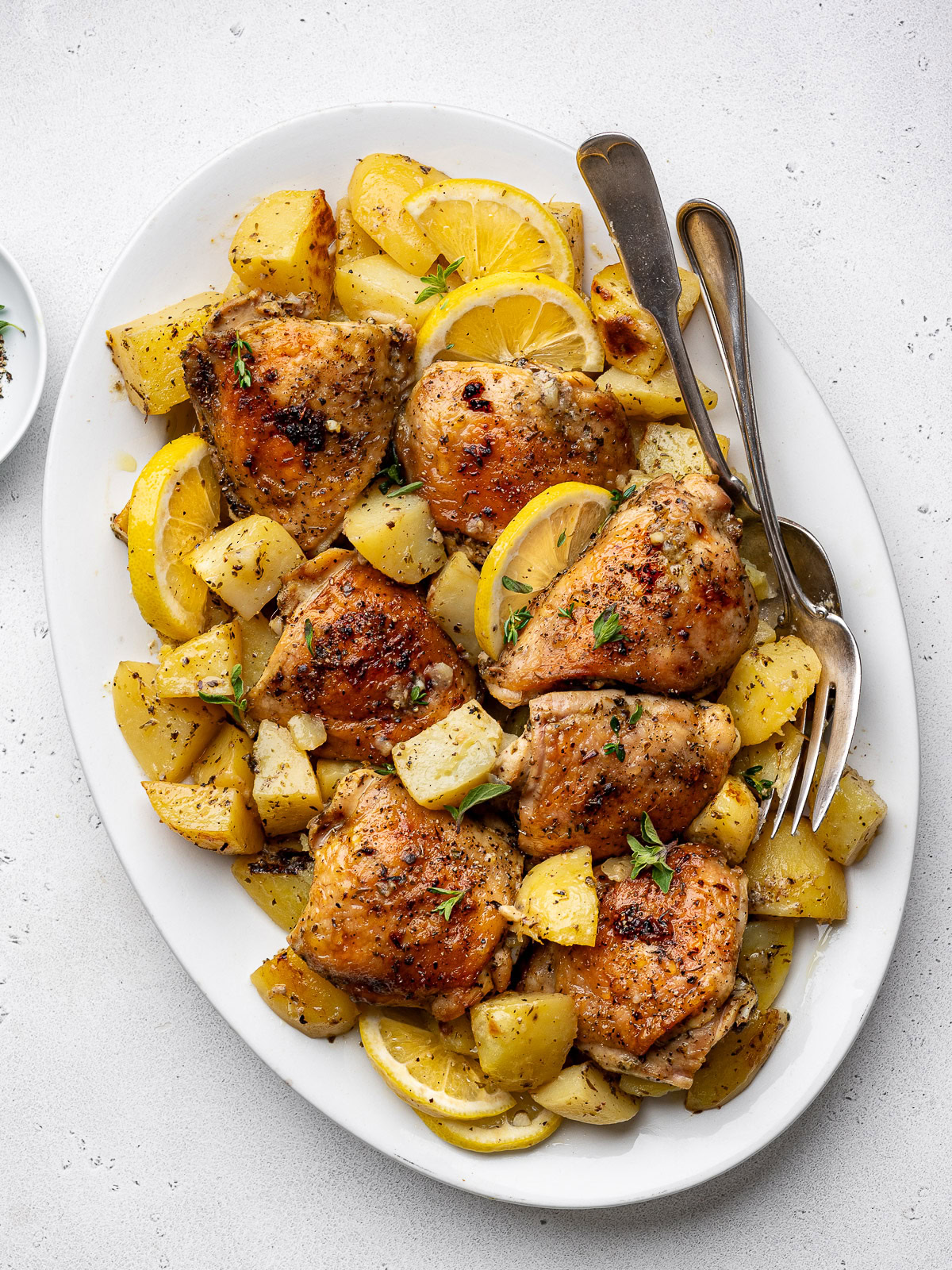 baked Greek chicken and potatoes in a white casserole dish garnished with lemon slices and fresh oregano.