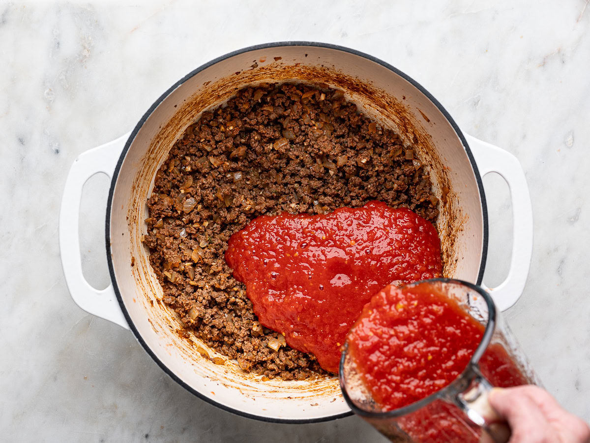 pouring crushed tomatoes over ground meat mixture in pot