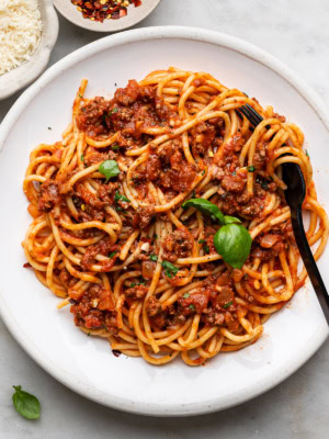 spaghetti and meat sauce served on a white plate with black fork and a small bowl of parmesan and chili flakes on the side.