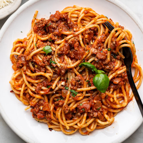 spaghetti and meat sauce served on a white plate with black fork and a small bowl of parmesan and chili flakes on the side.