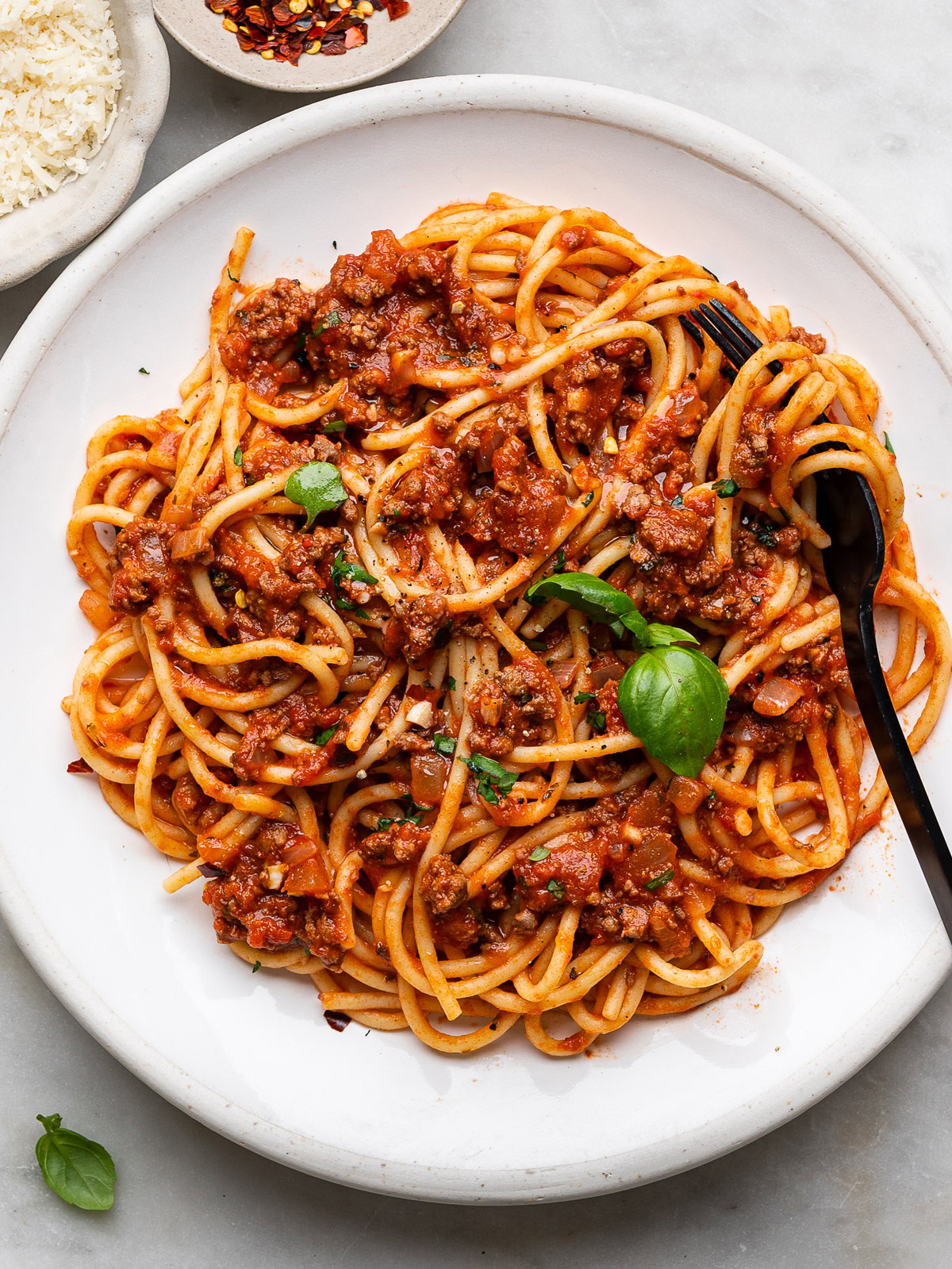 spaghetti and meat sauce served on a white plate with black fork and a small bowl of parmesan and chili flakes on the side. 