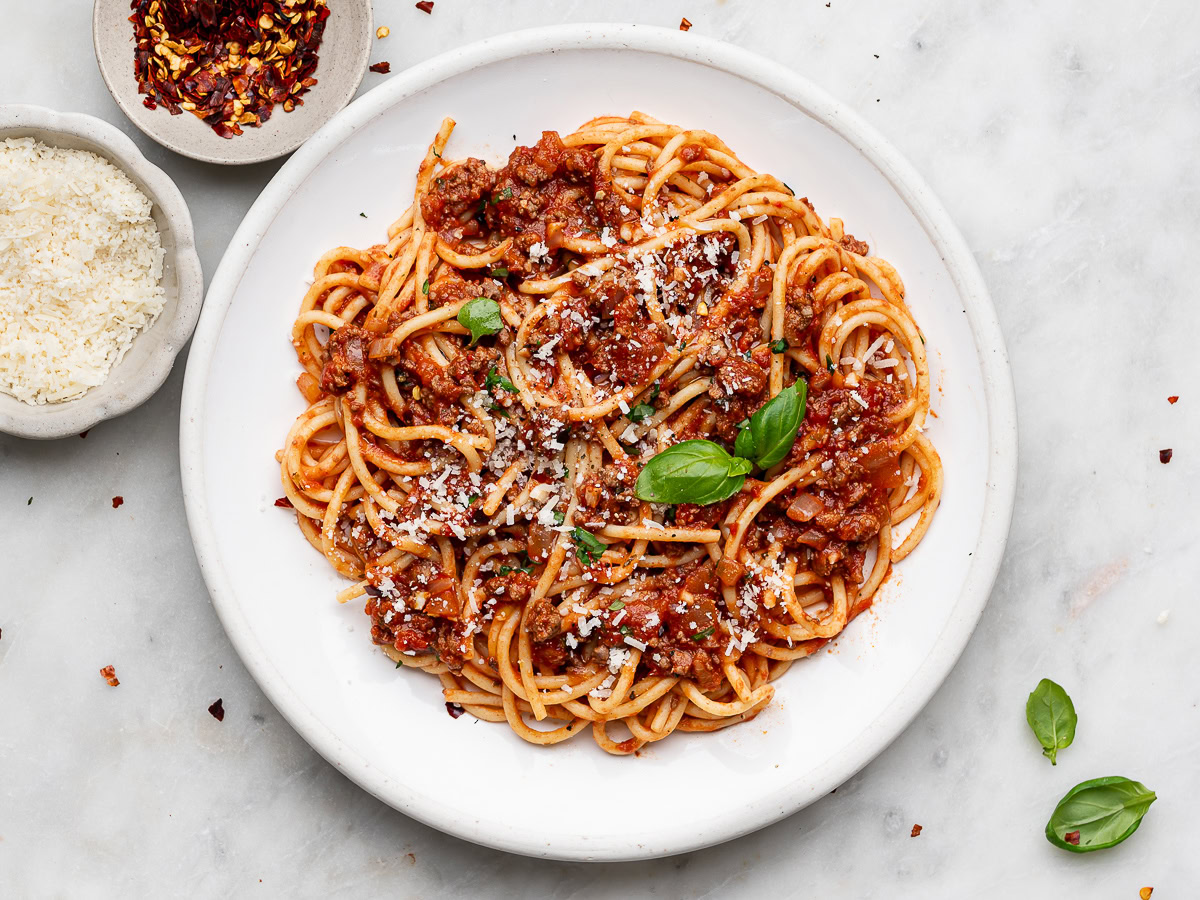 spaghetti and meat sauce served on a white plate with black fork and a small bowl of parmesan and chili flakes on the side.