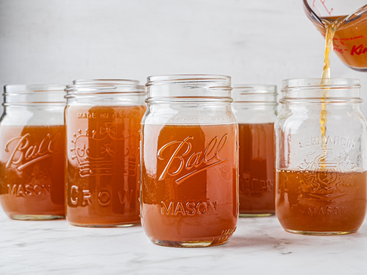 bone broth being poured into a mason jar with a measuring cup alongside other filled mason jars