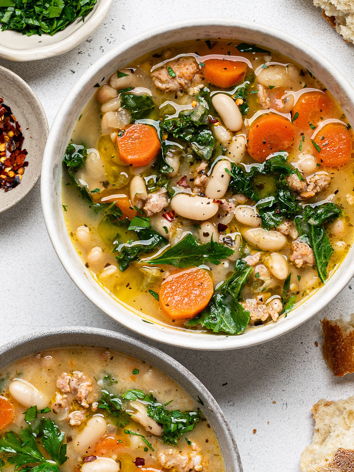 two bowls served with kale and white bean sausage soup with bowls of chopped parsley and red pepper flakes on the side and crusty bread