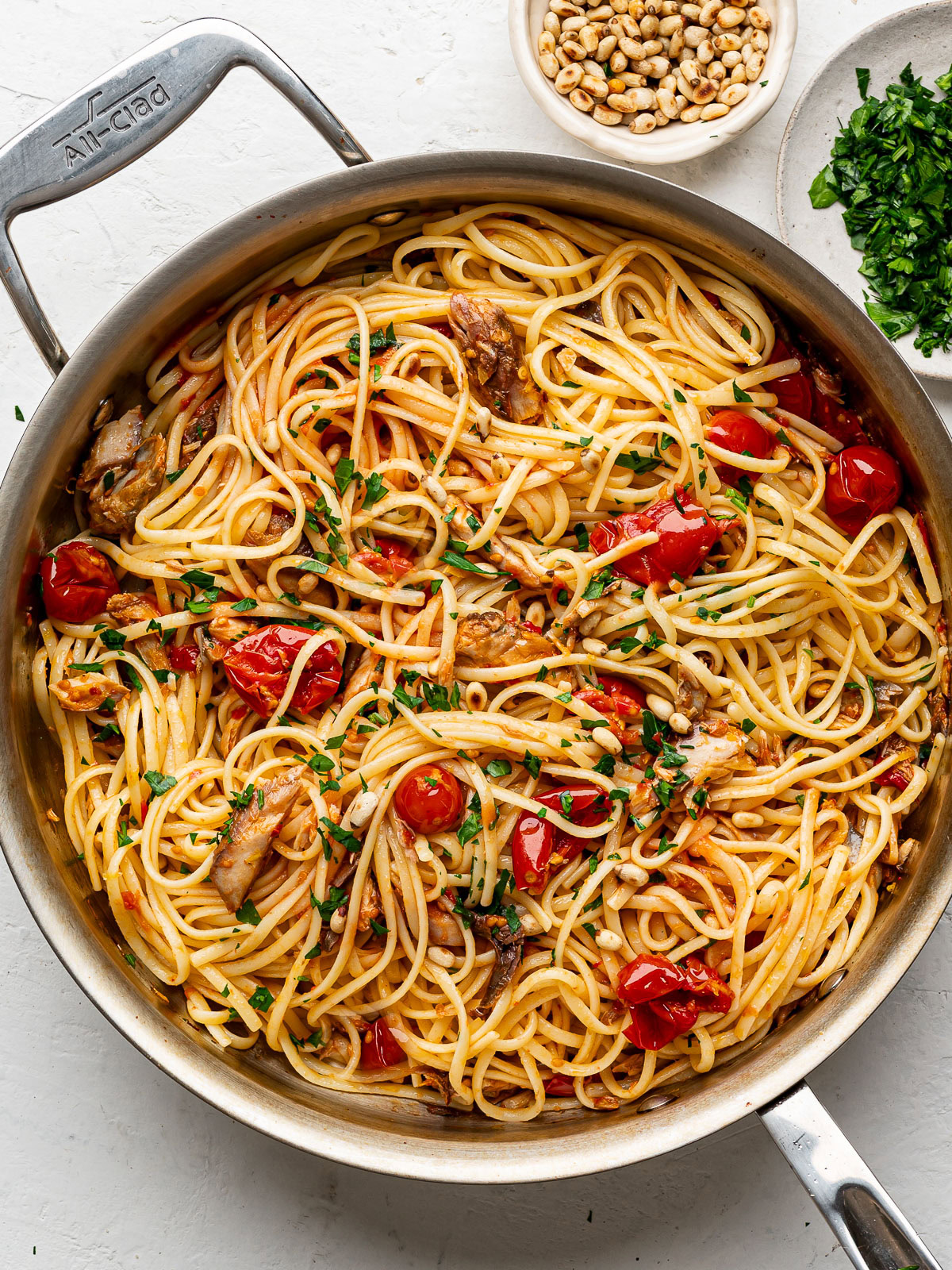 close up of mackerel pasta mixture in large stainless steel pan garnished with pine nuts and chopped parsley