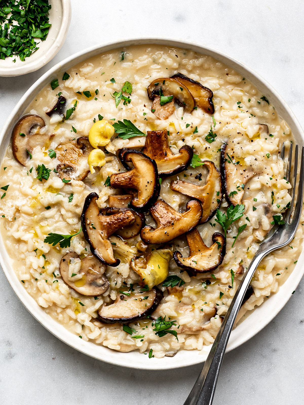 Mushroom risotto served in white plate topped with extra mushrooms and a small bowl of chopped parsley on the side