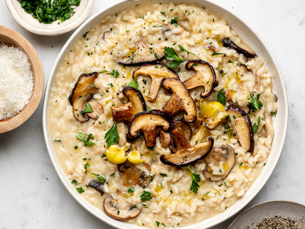 Mushroom risotto served in a white plate topped with extra mushrooms and a small bowl of chopped parsley and parmesan cheese on the side