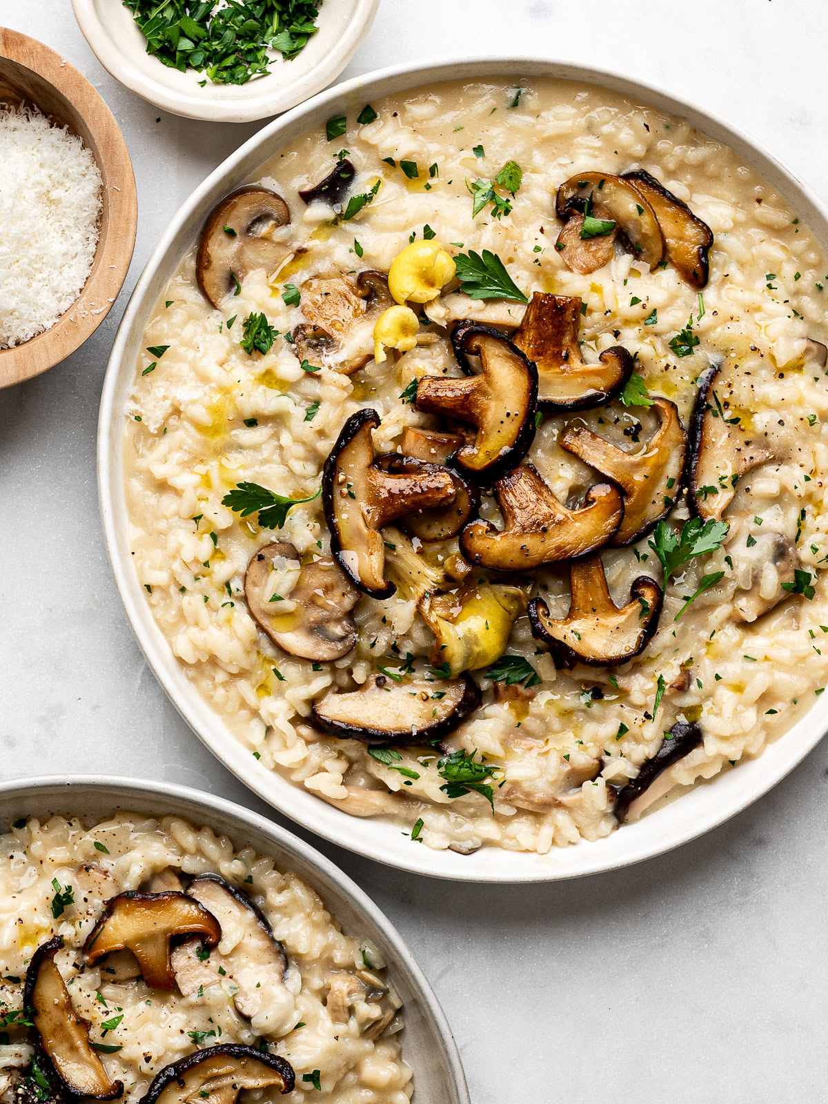 Mushroom risotto served in two white plates topped with extra mushrooms and a small bowl of chopped parsley and parmesan cheese on the side