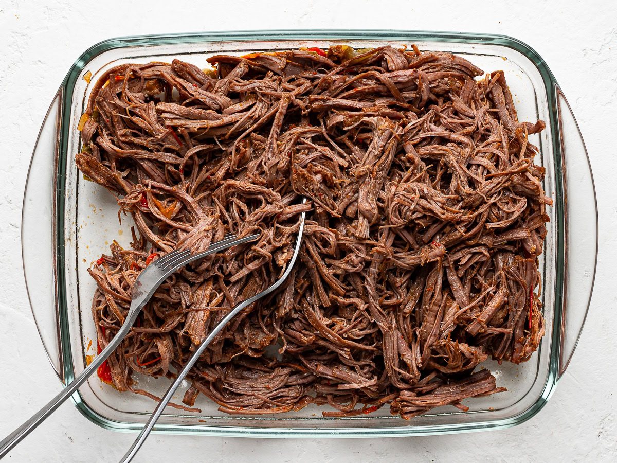 shredded flank steak in a glass rectangular bowl with two forks