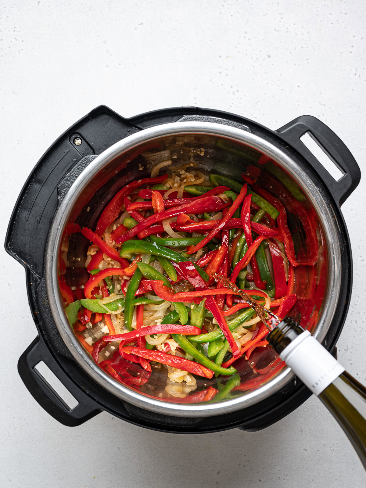 a bottle of wine shown pouring white wine over sautéed onion and bell peppers