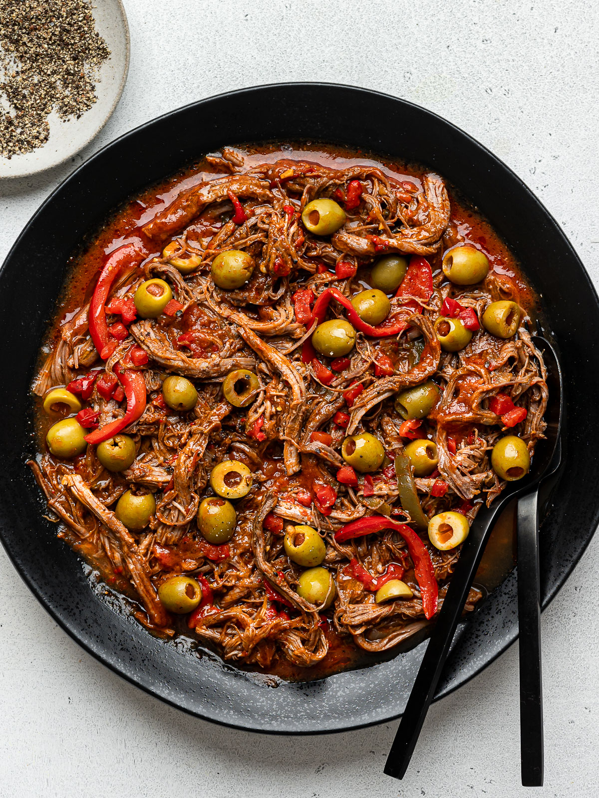 Ropa Vieja garnished with pimentos and olives served on a black round platter with black serving fork and spoon