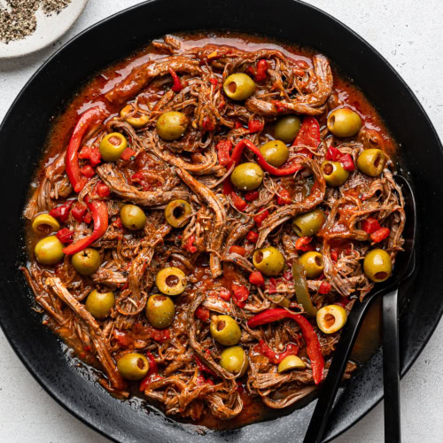 Ropa Vieja garnished with pimentos and olives served on a black round platter with black serving fork and spoon