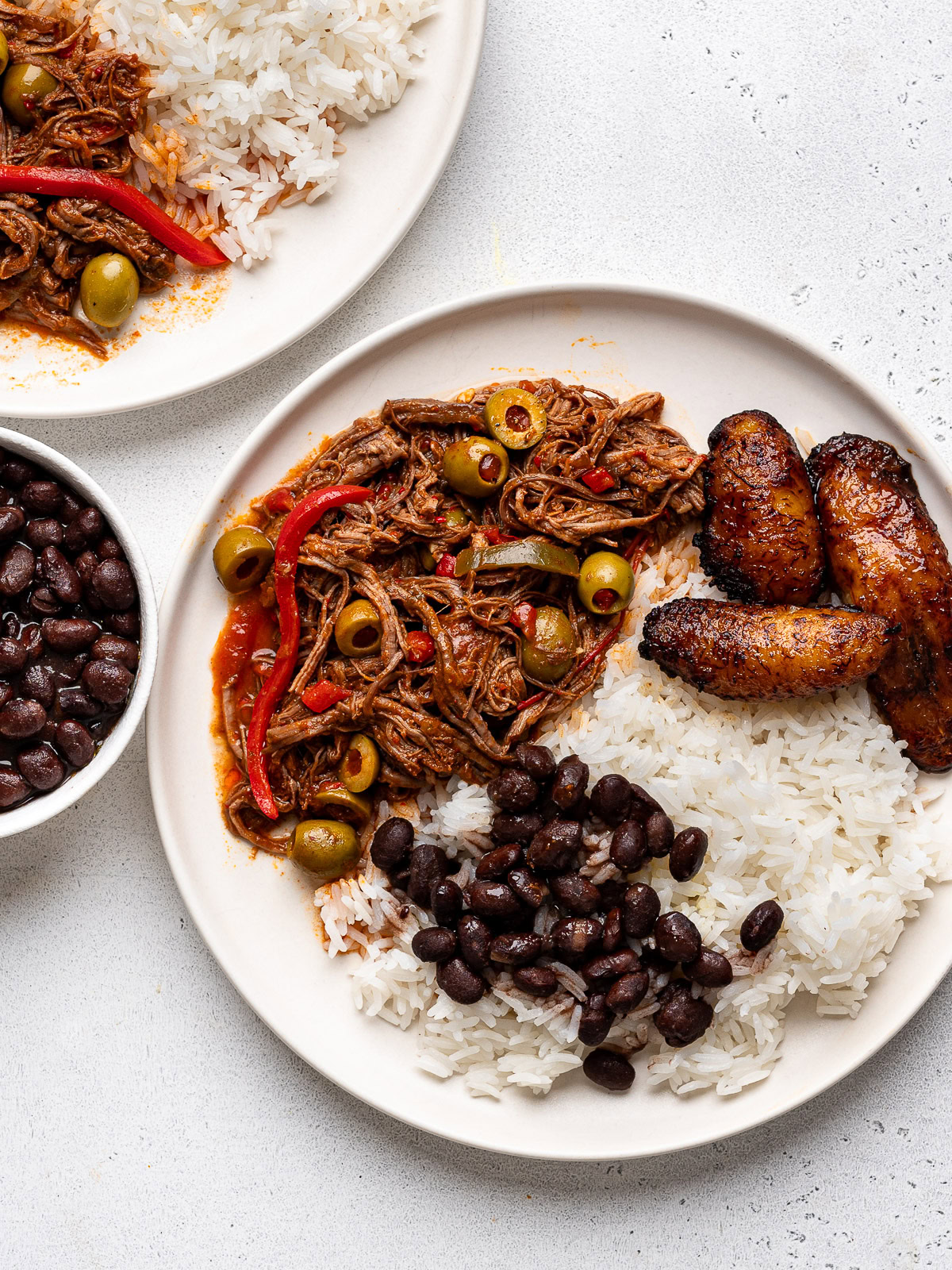 ropa vieja served on two white plates with white rice, black beans and fried plantains with a small bowl filled with black beans on the side