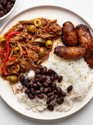 close up of ropa vieja served on white plates with white rice, black beans and fried plantains with a small bowl filled with black beans on the side