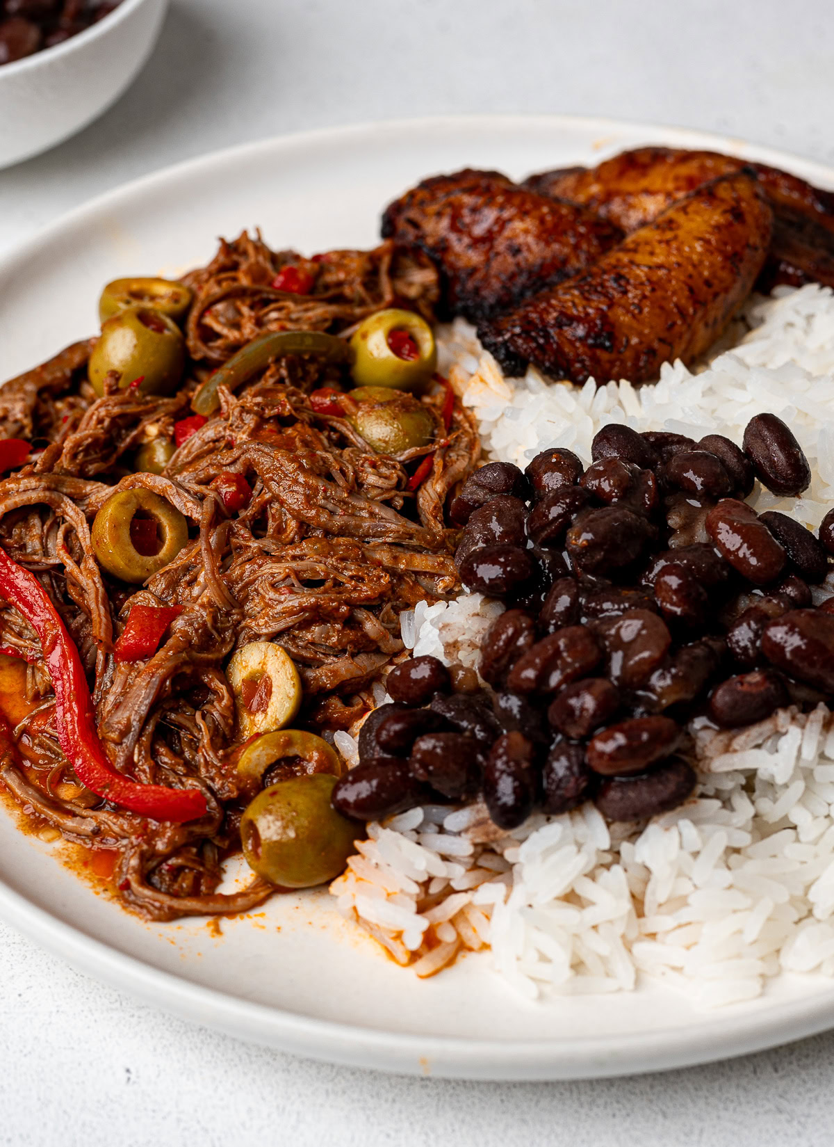 closeup image of ropa vieja served on a white plate with white rice, black beans and fried plantains