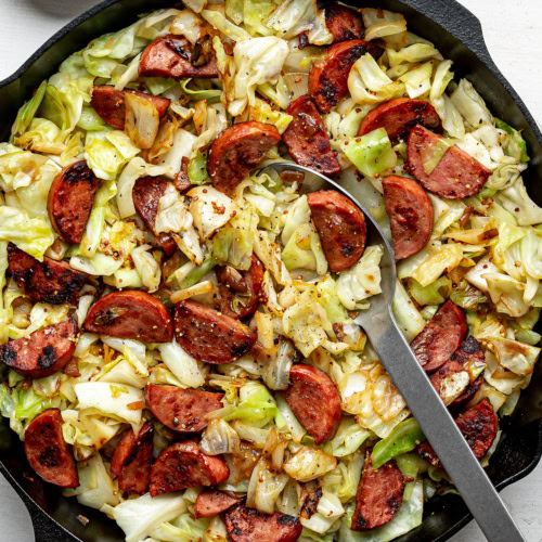 the finished dish of Sautéed cabbage and sausage in a black skillet with a large serving spoon
