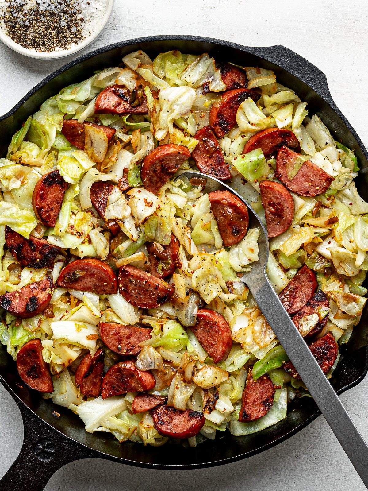 The finished dish of Sautéed cabbage and sausage in a black skillet with a large serving spoon. 