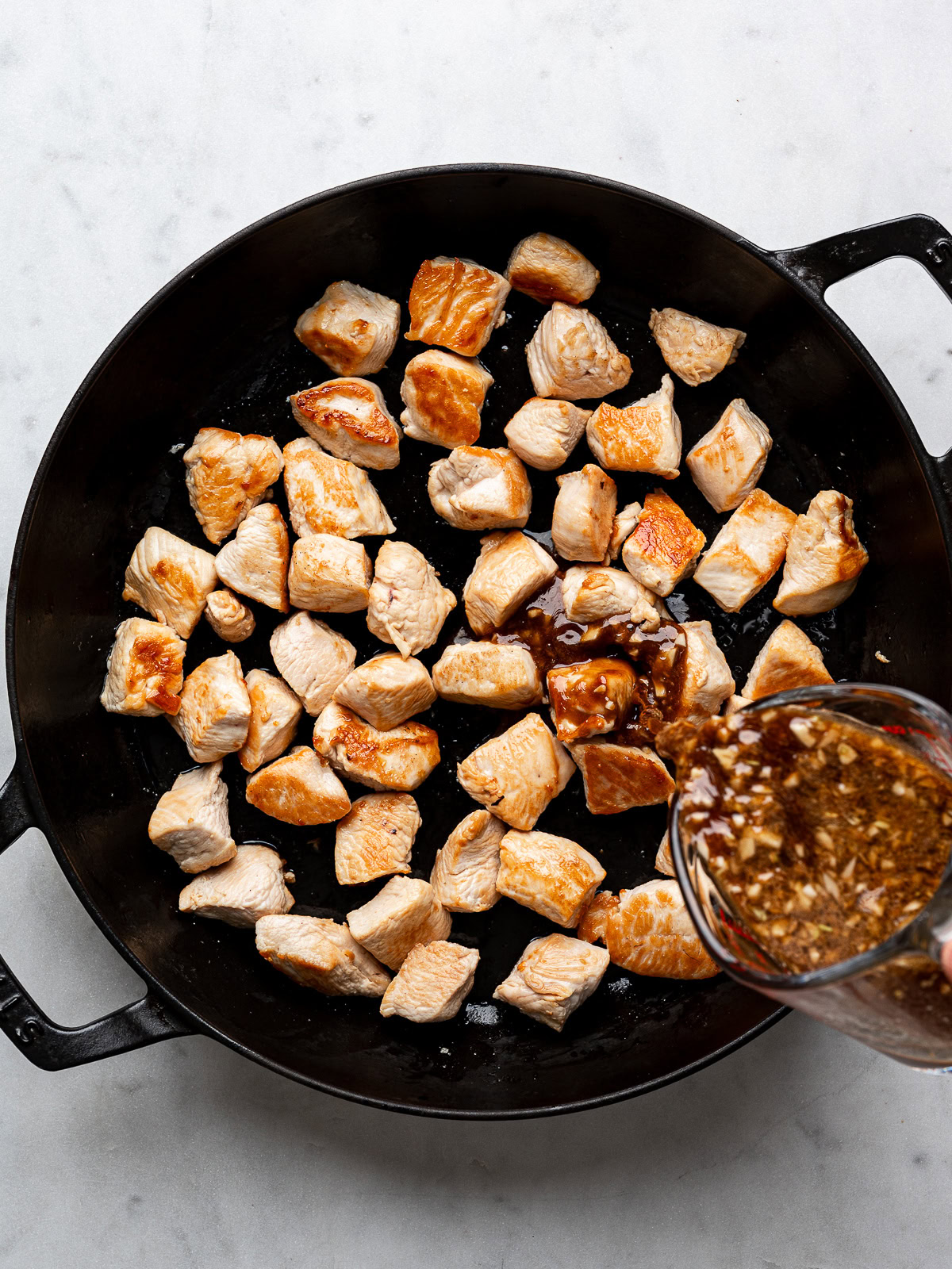 Pouring teriyaki sauce over cooked chicken in black skillet.