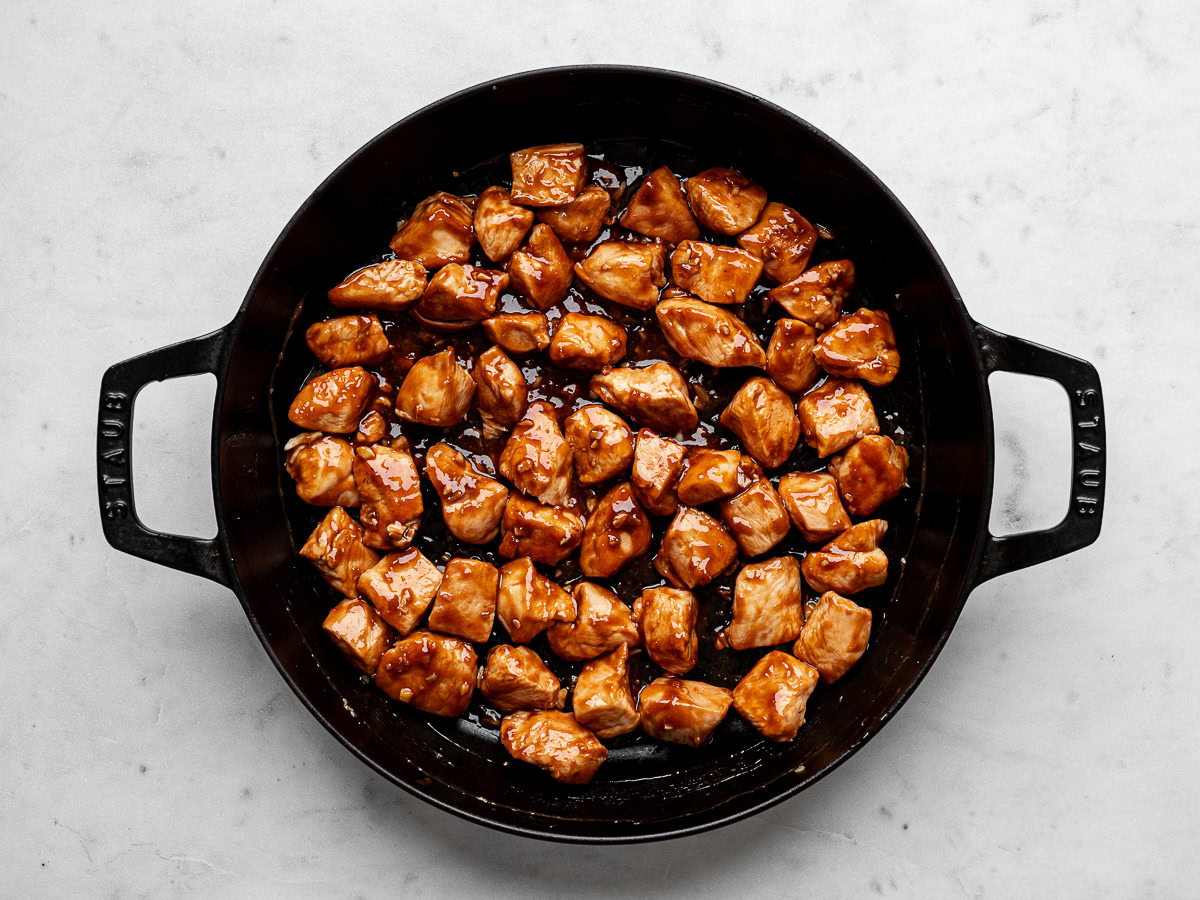 Cubed cooked chicken pieces coated with teriyaki sauce in black skillet.