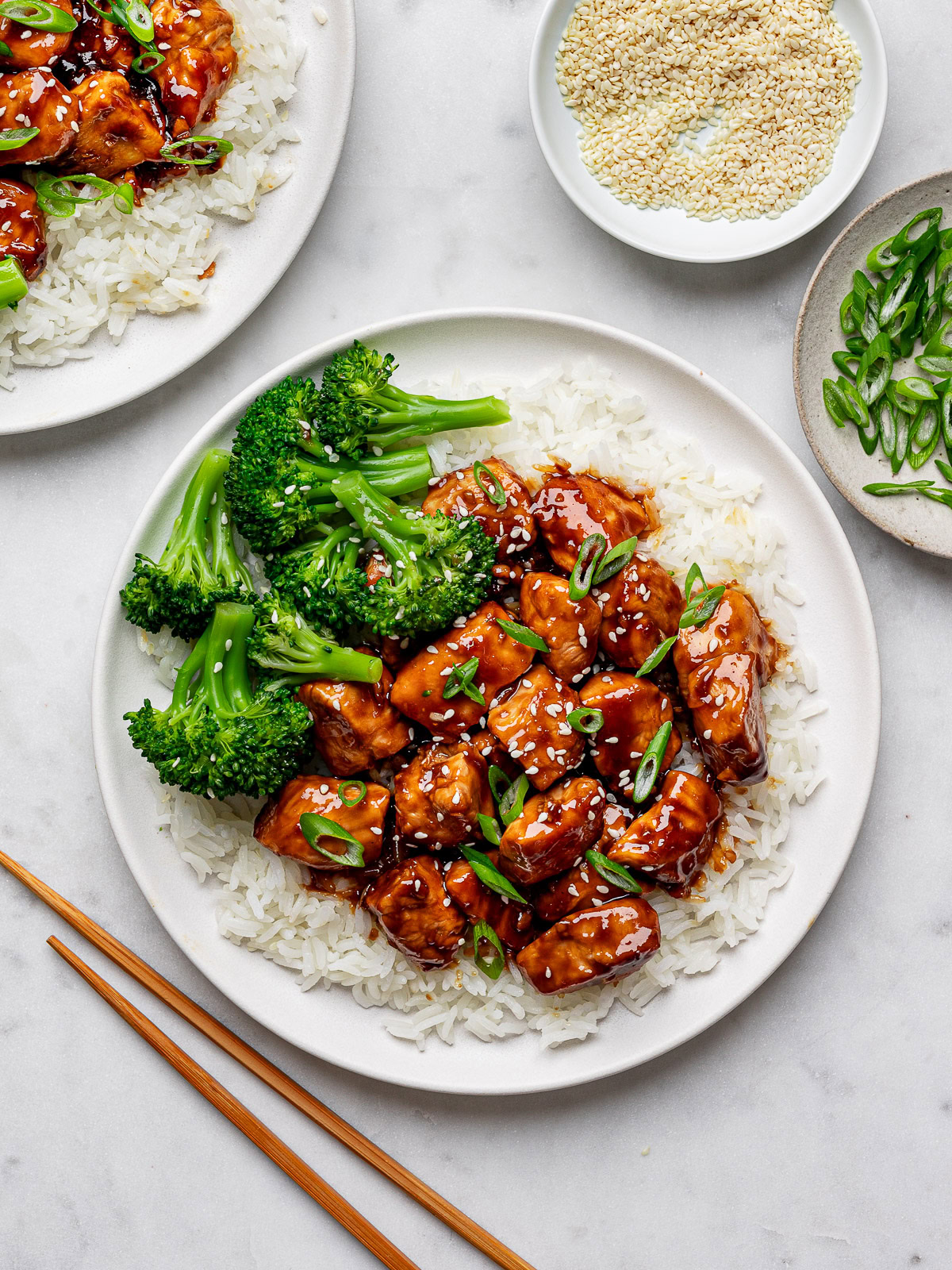 Two white plates with Teriyaki chicken served over fluffy white rice with steamed broccoli on the side and small pinch bowls with sesame seeds and sliced green onions.