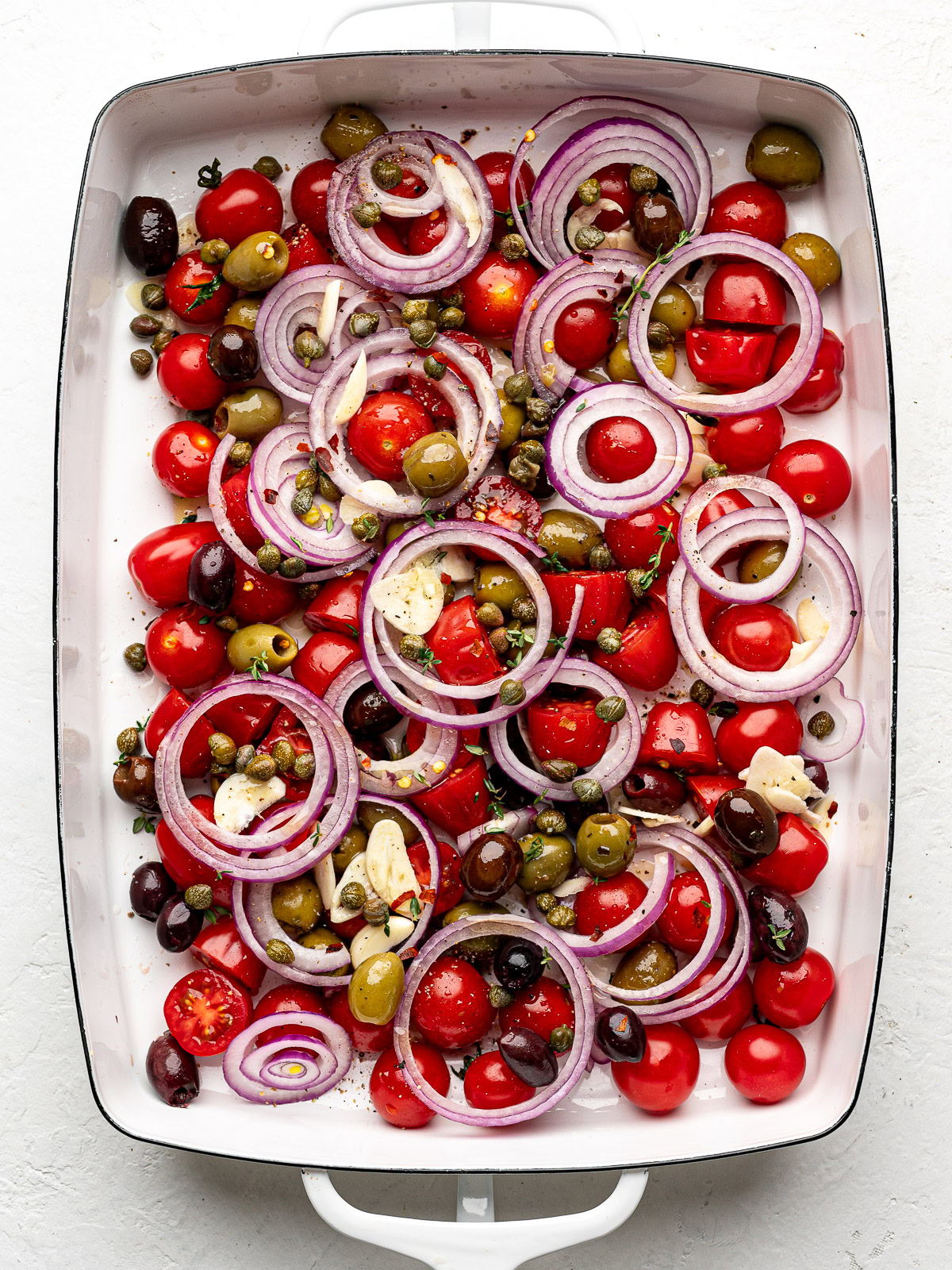 Cherry tomato mixture with onion rings and olives in baking dish before baking.