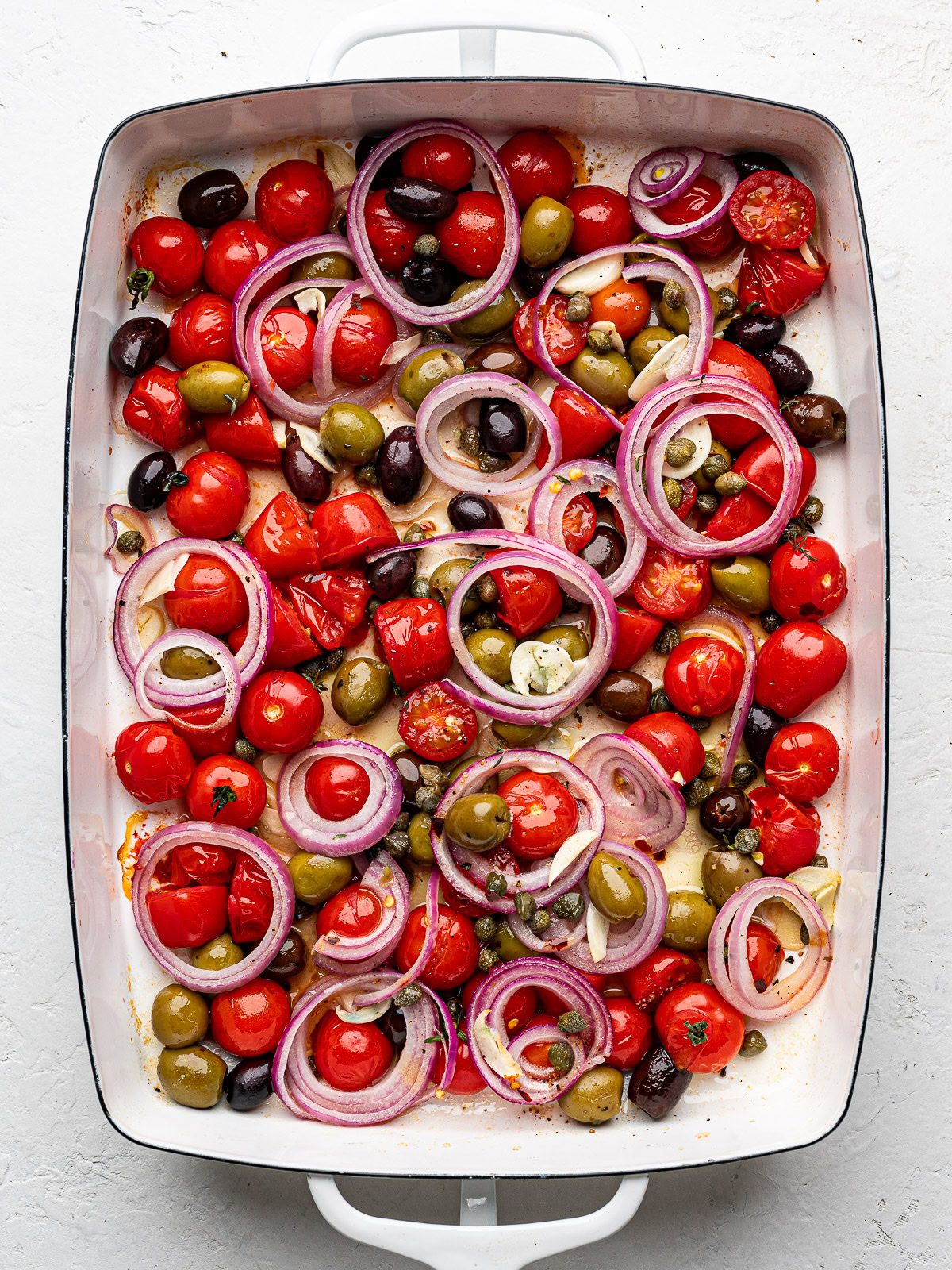 Baked burst cherry tomato mixture with onion rings and olives in baking dish.